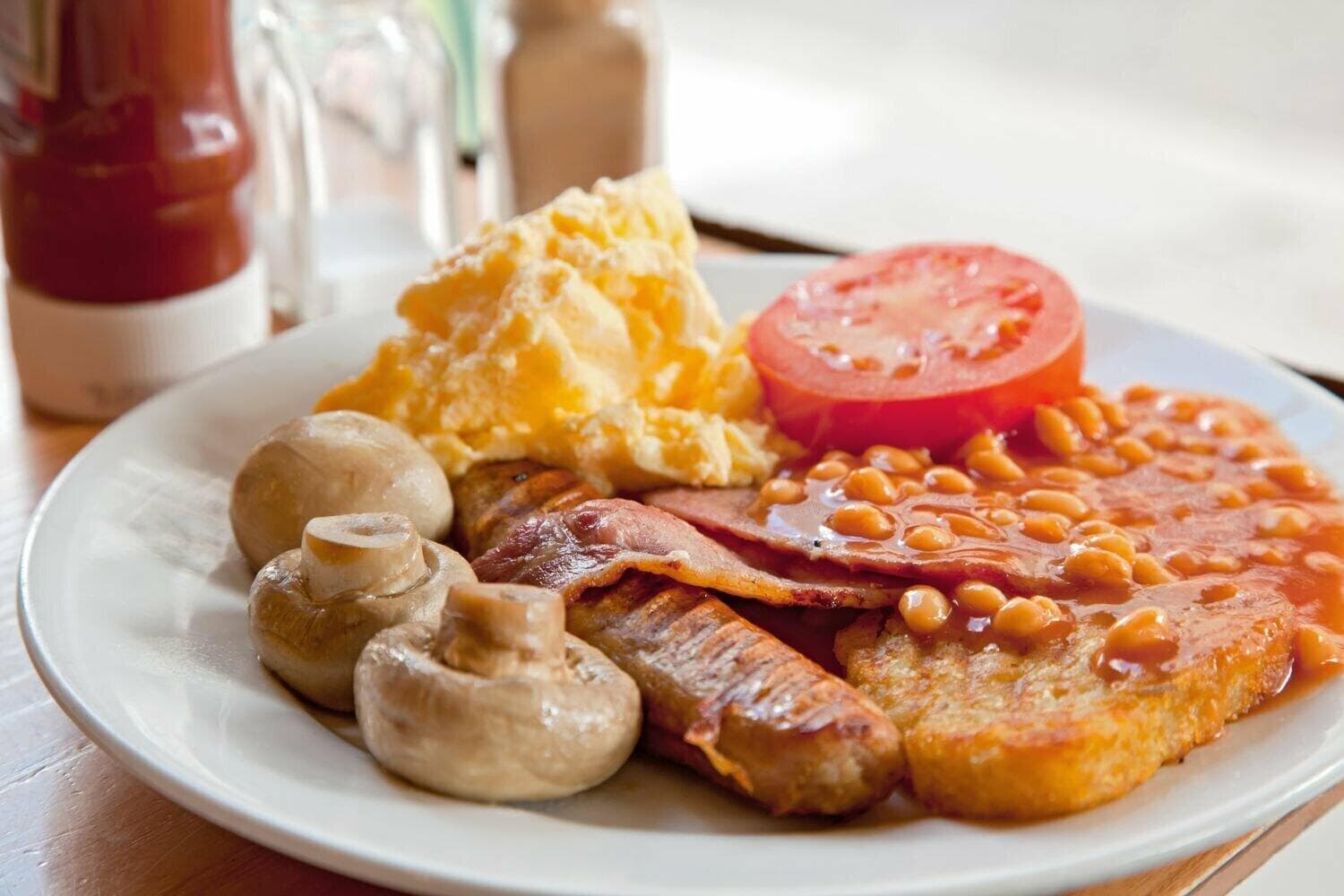A cooked breakfast of scrambled eggs, bacon, sausage, beans, mushrooms, tomato and a hash brown