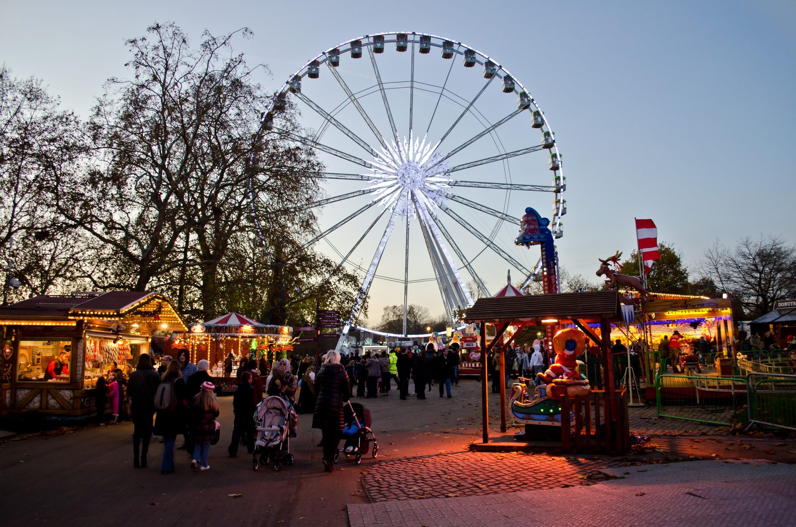 Hyde Park Winter Wonderland at night, with fairground rides and the Giant Wheel lit above the trees